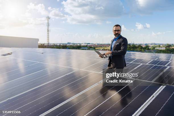 portrait of businessman holding laptop with standing on rooftop which install photovoltaic panel system. - cleantech stock pictures, royalty-free photos & images