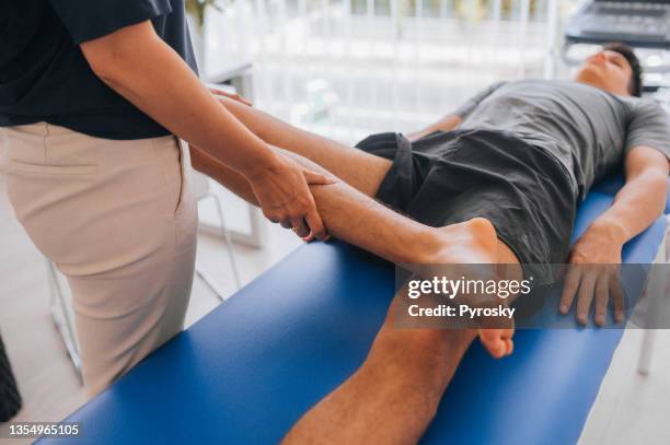 female physiotherapist bending the knee of an injured male patient lying on a massage bed in the clinic - osteopath stock pictures, royalty-free photos & images