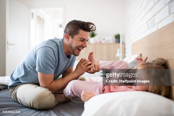 happy young father having fun with little daughter together at home. happy young man sitting on bed and tickling his small cute daughter on foot indoors at home. - tickling stock-fotos und bilder