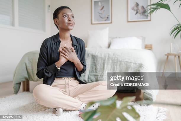 black woman with hand on chest showing gratitude while praying sitting on the floor in her bedroom. - danke stock-fotos und bilder