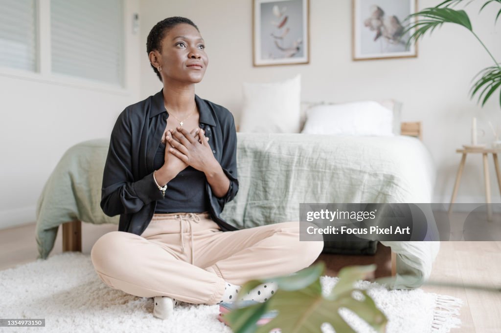 Black woman with hand on chest showing gratitude while praying sitting on the floor in her bedroom.