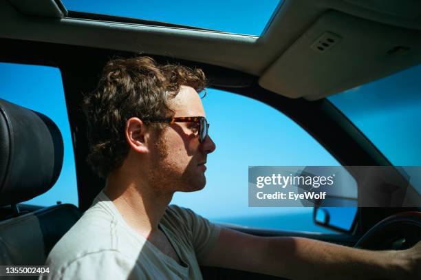 young man driving along coast in car the sunroof, wearing sunglasses - car sunroof stockfoto's en -beelden