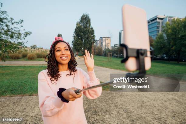 woman waving hand while vlogging through smart phone at park - selfie stick stock pictures, royalty-free photos & images