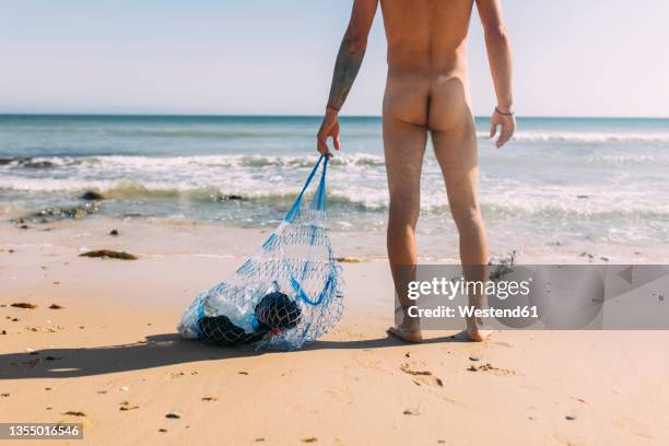 naked volunteer collecting plastic pollution in mesh bag at beach - beach bum foto e immagini stock