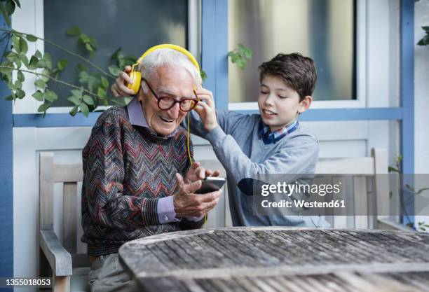 boy adjusting grandfather's headphones on bench - abuelos fotografías e imágenes de stock