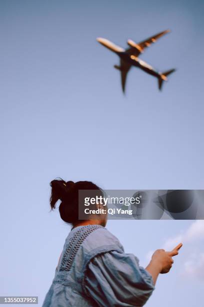 young woman holding smartphone looking up at airplane - evasion fiscale stock pictures, royalty-free photos & images