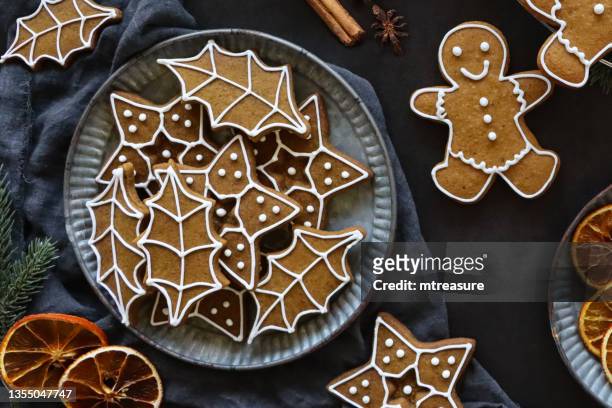 close-up image of batch of freshly baked, homemade, gingerbread holly leaf and star biscuits decorated with white royal icing on a plate, grey muslin, cinnamon stick, dried orange citrus slices, black background, elevated view - cookie studio stock pictures, royalty-free photos & images