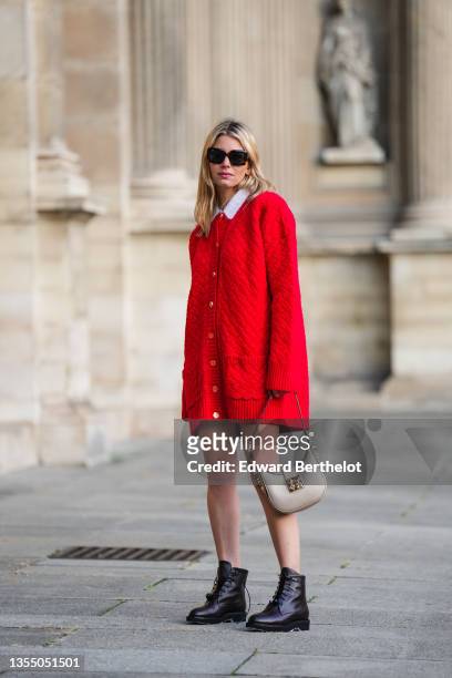 Natalia Verza wears sunglasses, a white wool t-shirt, a red shiny braided embossed pattern wool oversized cardigan with flower pattern buttons, a...