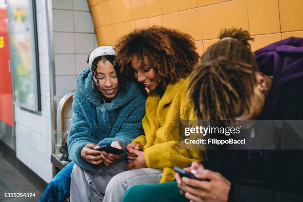 tres amigos esperando el tren en una estación de metro, mirando teléfonos inteligentes - london underground fotografías e imágenes de stock