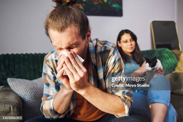 cropped shot of a handsome young man blowing his nose at home with his girlfriend in the background - allergie stockfoto's en -beelden