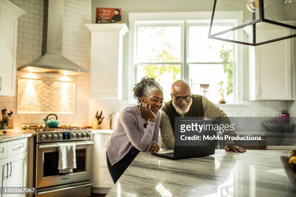 senior couple using laptop in kitchen of suburban home - pensioen thema stockfoto's en -beelden