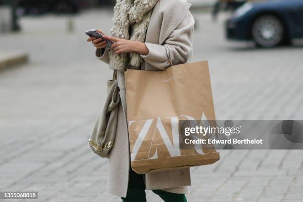 Passerby wears a beige with embroidered pompom wool scarf, a beige long wool coat, a beige with gold sequins handbag, dark green sport pants, a brown...