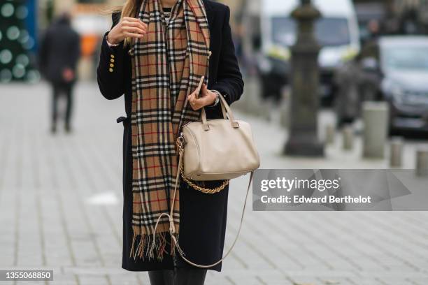 Passerby wears a black long wool coat, a brown / red / brown checkered print pattern fringed scarf from Burberry, a beige shiny leather handbag,...