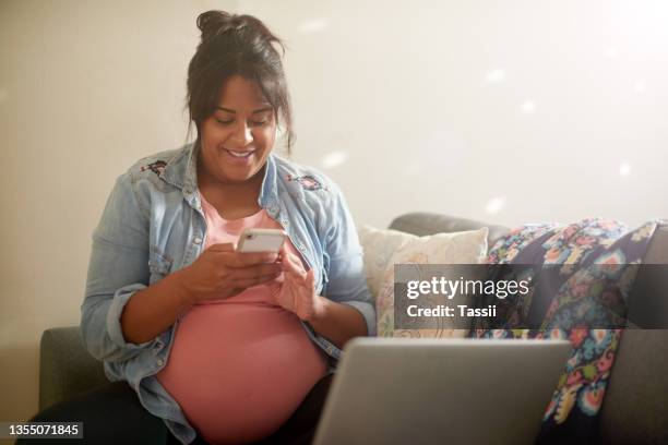 cropped shot of an attractive young pregnant woman using her cellphone and laptop while sitting in her living room at home - indian mother stockfoto's en -beelden