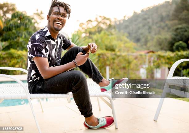 laughing young man sitting on a deck chair and texting on his phone - dreadlocks bildbanksfoton och bilder