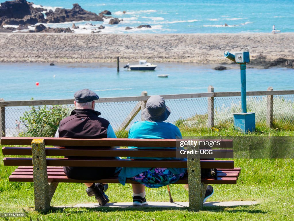 Old man and woman sat on a public bench looking out to sea, Bude