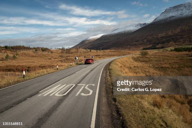 road with a slow warning sign on the tarmac across majestic autumn coloured mountains partially covered in snow in the glencoe valley, highlands of scotland, united kingdom, with a red car driving under a blue sky with clouds - bollards stock pictures, royalty-free photos & images