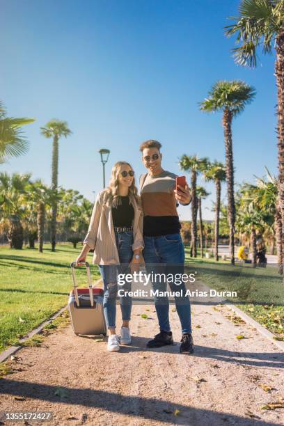 multiracial couple walking through a resort with their luggage taking a selfie - felicidad duran fotografías e imágenes de stock