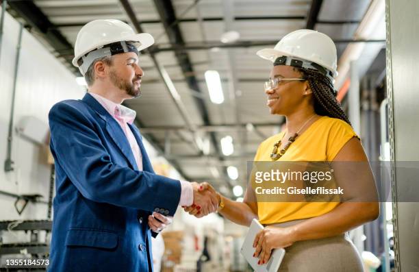 two smiling diverse businesspeople shaking hands in a large warehouse - business relationship stock pictures, royalty-free photos & images
