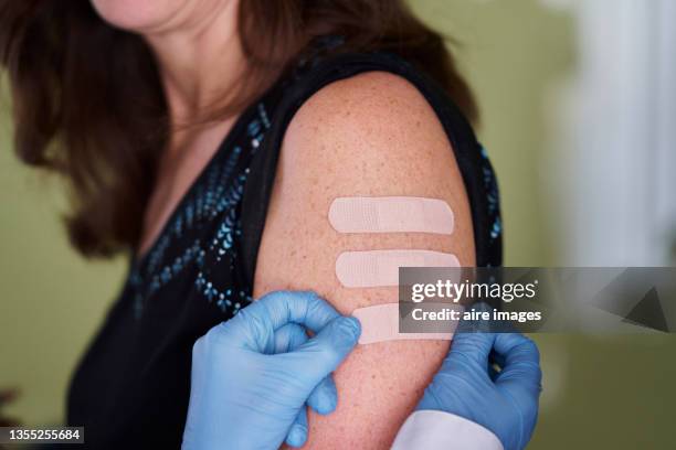close-up of rear view of a young female doctor wearing gloves applying a third adhesive bandage to a young caucasian woman after injecting the vaccine dose against a blurred background - 3 shot fotografías e imágenes de stock