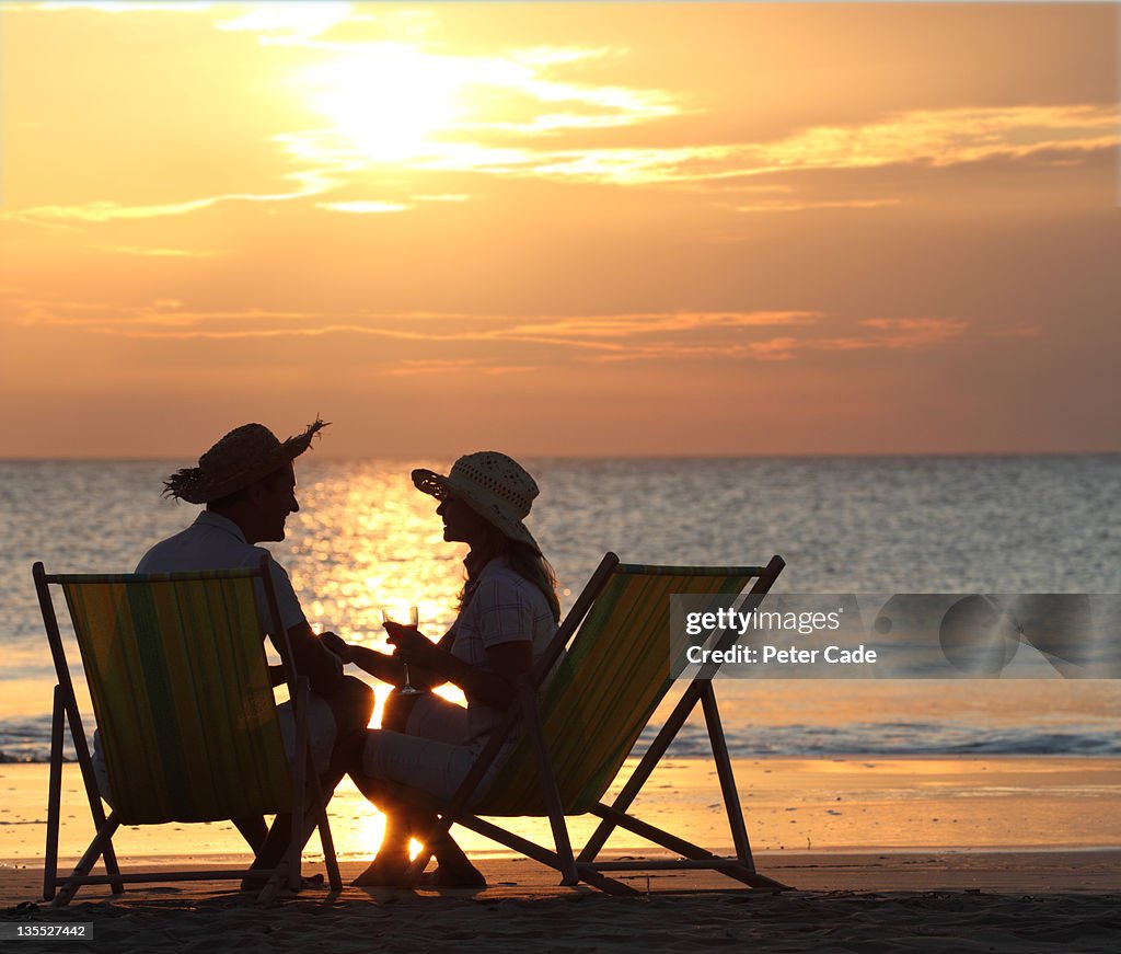 Couple sat in deckchairs on beach at sunset