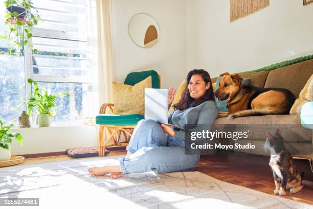 full length shot of an attractive young woman sitting in her living room with her pets and using her laptop - cat owner stockfoto's en -beelden