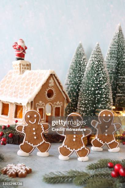 image of three gingerbread men skating in front of homemade, gingerbread house decorated with white royal icing displayed in snowy conifer forest scene, santa claus, model fir trees, icing sugar snow, snowy blue background - cookie monster stockfoto's en -beelden