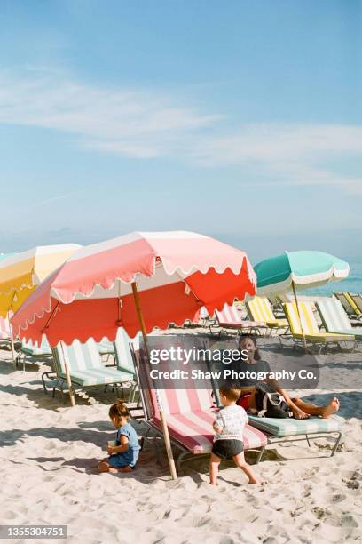 mom sitting on a beach chair on the beach looking at the camera while kids play around her with bright beautiful umbrellas around her and sand beneath her and a bright blue sky in miami beach florida - miami beach fotografías e imágenes de stock