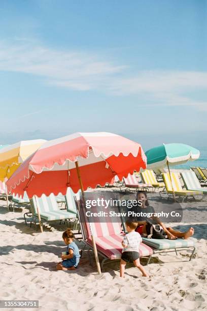 mom sitting on a beach chair on the beach looking at the camera while kids play around her with bright beautiful umbrellas around her and sand beneath her and a bright blue sky in miami beach florida - florida beaches 個照片及圖片檔