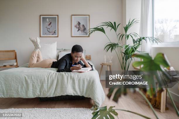 black woman writing in a notebook while laying on a comfortable bed at home. - bedroom stockfoto's en -beelden