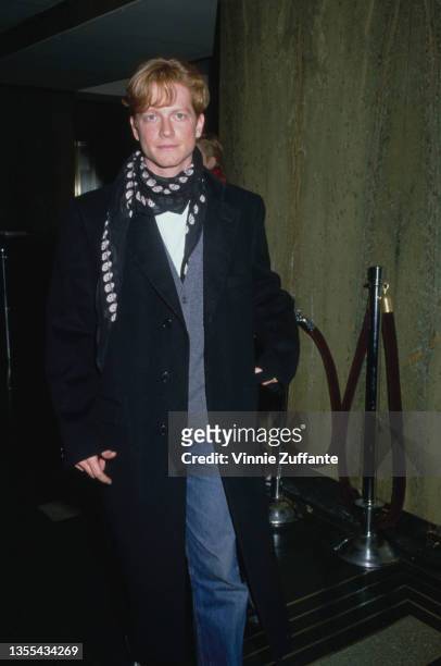 American actor Eric Stoltz, wearing a grey cardigan and jeans beneath a long black coat and a spotted scarf around his neck, circa 1995.