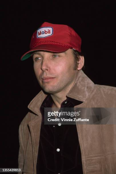 American singer-songwriter and guitarist Dwight Yoakam, wearing a tan suede jacket over a black shirt with a red 'Mobil' baseball cap, attends the...