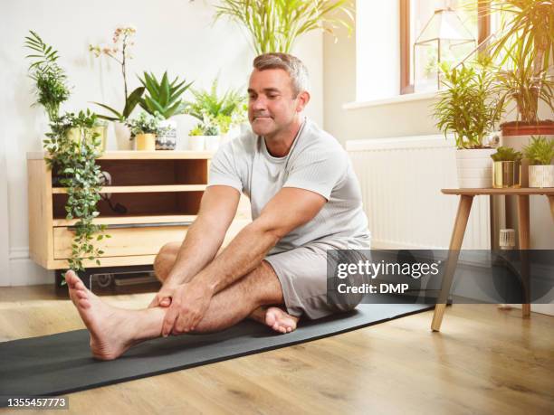 shot of a mature man stretching his legs while practicing yoga at home - yoga mat stock pictures, royalty-free photos & images