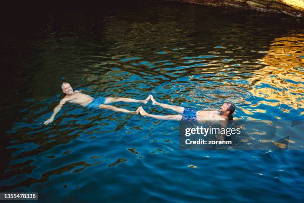 couple gay d’âge moyen heureux flottant sur l’eau, touchant avec les pieds - gay men swimwear photos et images de collection