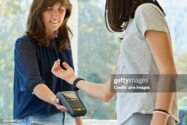 rear view of a teenager girl paying using her smart watch while the female cashier holding the pos in a clothing store of the mall - smartwatch pay stock pictures, royalty-free photos & images