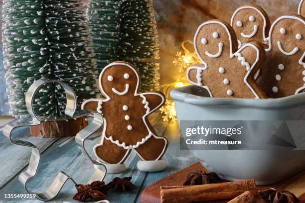 close-up image of tobogganing scene with batch of homemade, gingerbread men biscuits, iced with white, royal icing, sat in ceramic cake pan toboggan, cookie cutter, model fir trees and illuminated fairy lights on blue wood grain background - cookie cutter stock pictures, royalty-free photos & images