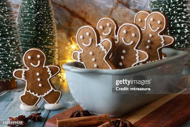 image of tobogganing scene with batch of homemade, gingerbread men biscuits, iced with white, royal icing, sat in ceramic cake pan toboggan, cinnamon sticks, star anise and model fir trees and illuminated fairy lights on blue wood grain background - pastry cutter stockfoto's en -beelden