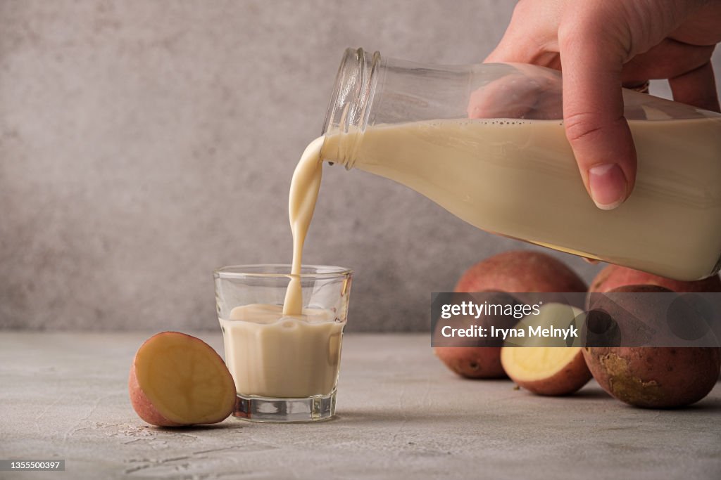 Woman is pouring Potato milk alternative non dairy drink in glass from bottle on gray background. Healthy vegetarian and vegan drink concept