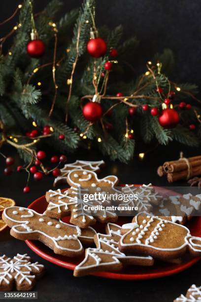 close-up image of plate of gingerbread men and snowflake cookies with white royal icing, star anise, bundle of cinnamon sticks, dried citrus orange fruit slices, mini christmas tree and red berries, illuminated fairy lights on black background - star fruit stockfoto's en -beelden