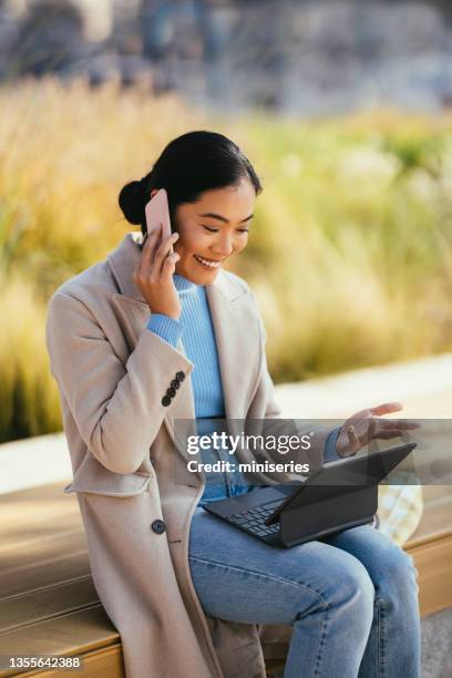 smiling asian woman sitting on a bench and talking on a phone while holding a digital tablet on her lap - surprised woman looking at tablet stock pictures, royalty-free photos & images