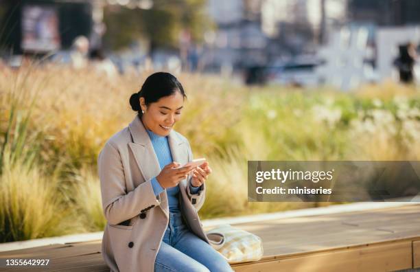 smiling asian woman sitting outside and looking at her phone - sincere chat stockfoto's en -beelden