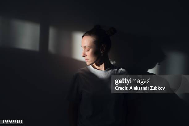 a lonely young sad calm girl or woman stands against a white wall, in a psychiatric clinic or at home, in the rays of sunlight, with her eyes closed. the concept of depression from illness and quarantine, fatigue and impotence. a copy of the space. - sombre stock-fotos und bilder