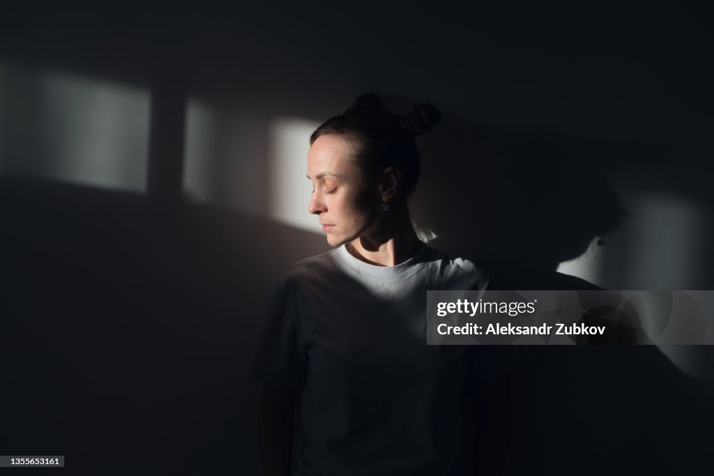 A lonely Young Sad Calm Girl Or Woman stands against a white wall, in a psychiatric clinic or at home, in the rays of sunlight, with her eyes closed. The Concept of Depression from Illness and Quarantine, Fatigue and Impotence. A copy of the space.