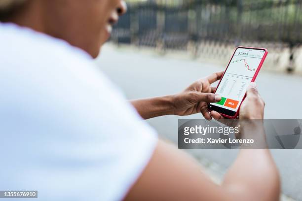 woman using an investing application on her mobile phone - african american woman with money stock-fotos und bilder
