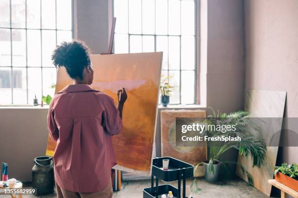 anonymous afro female artist wearing a pink shirt, painting on canvas in her home studio - watercolour orange and black stockfoto's en -beelden