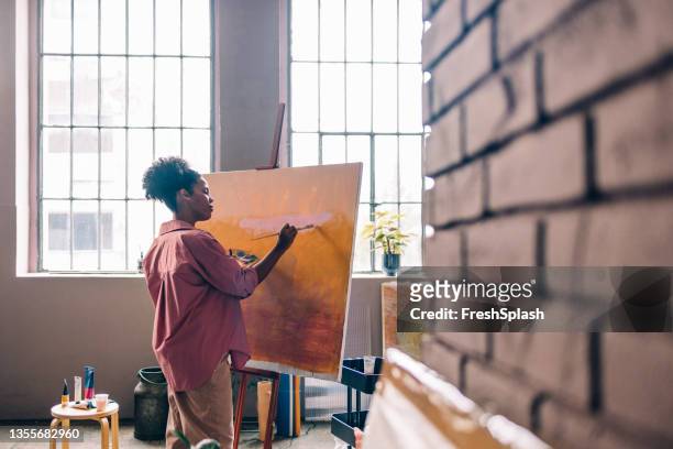 schöne afro-künstlerin trägt ein rosa hemd und malt auf leinwand in ihrem heimstudio - watercolour orange and black stock-fotos und bilder