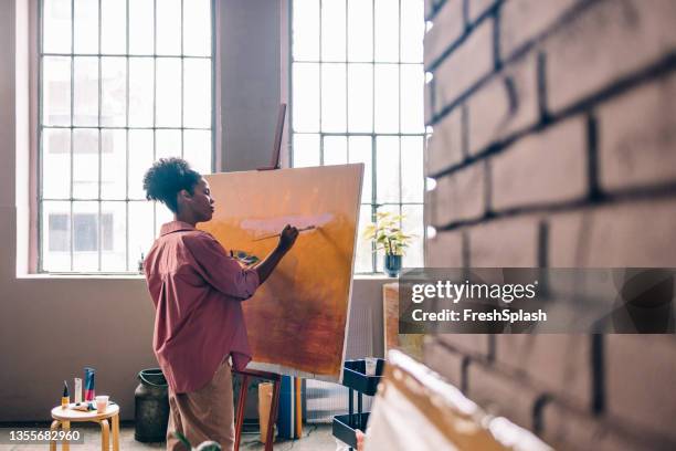 beautiful afro female artist wearing a pink shirt, painting on canvas in her home studio - painter artist 個照片及圖片檔