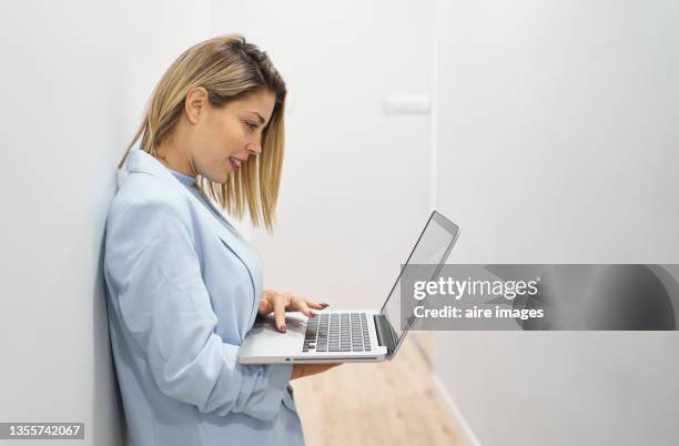 side view of blonde-haired woman standing in office hallway using laptop. woman in business attire smiling while video conferencing with business associates. - video wall fotografías e imágenes de stock