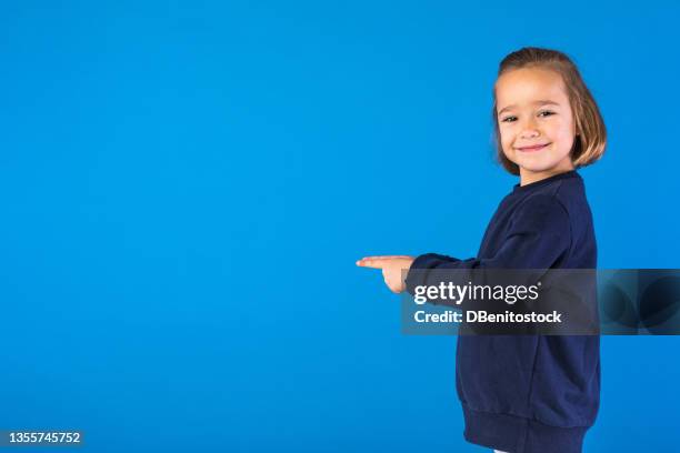 little blond hair girl wearing blue sweatshirt, pointing to the left with her hands and fingers, smiling, on light blue background. - child pointing stock pictures, royalty-free photos & images