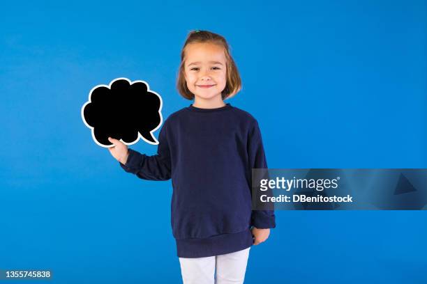 little blond-haired girl wearing blue sweatshirt, smiling, holding a black comic speech bubble in her hand, on light blue background. - small placard stock pictures, royalty-free photos & images